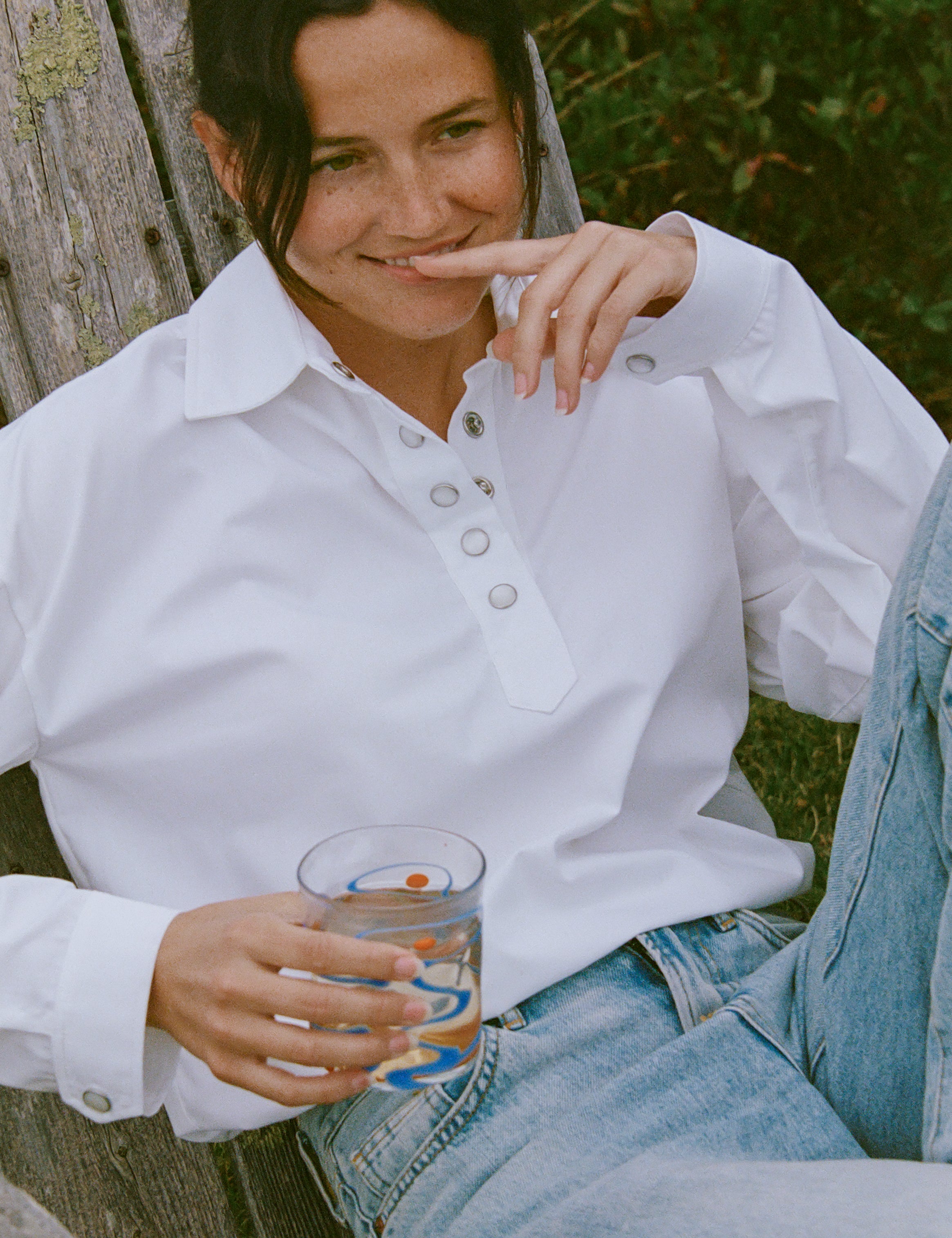 A woman sitting outdoors on a weathered wooden bench, wearing the darty popover in white and light blue jeans, holding a glass.