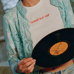 Close-up of the Going Out Top, white cropped tee with orange embroidered lettering, under a turquoise lace shirt with jeans