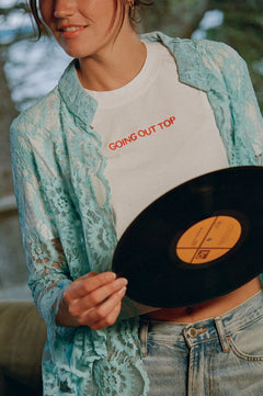 Close-up of the Going Out Top, white cropped tee with orange embroidered lettering, under a turquoise lace shirt with jeans