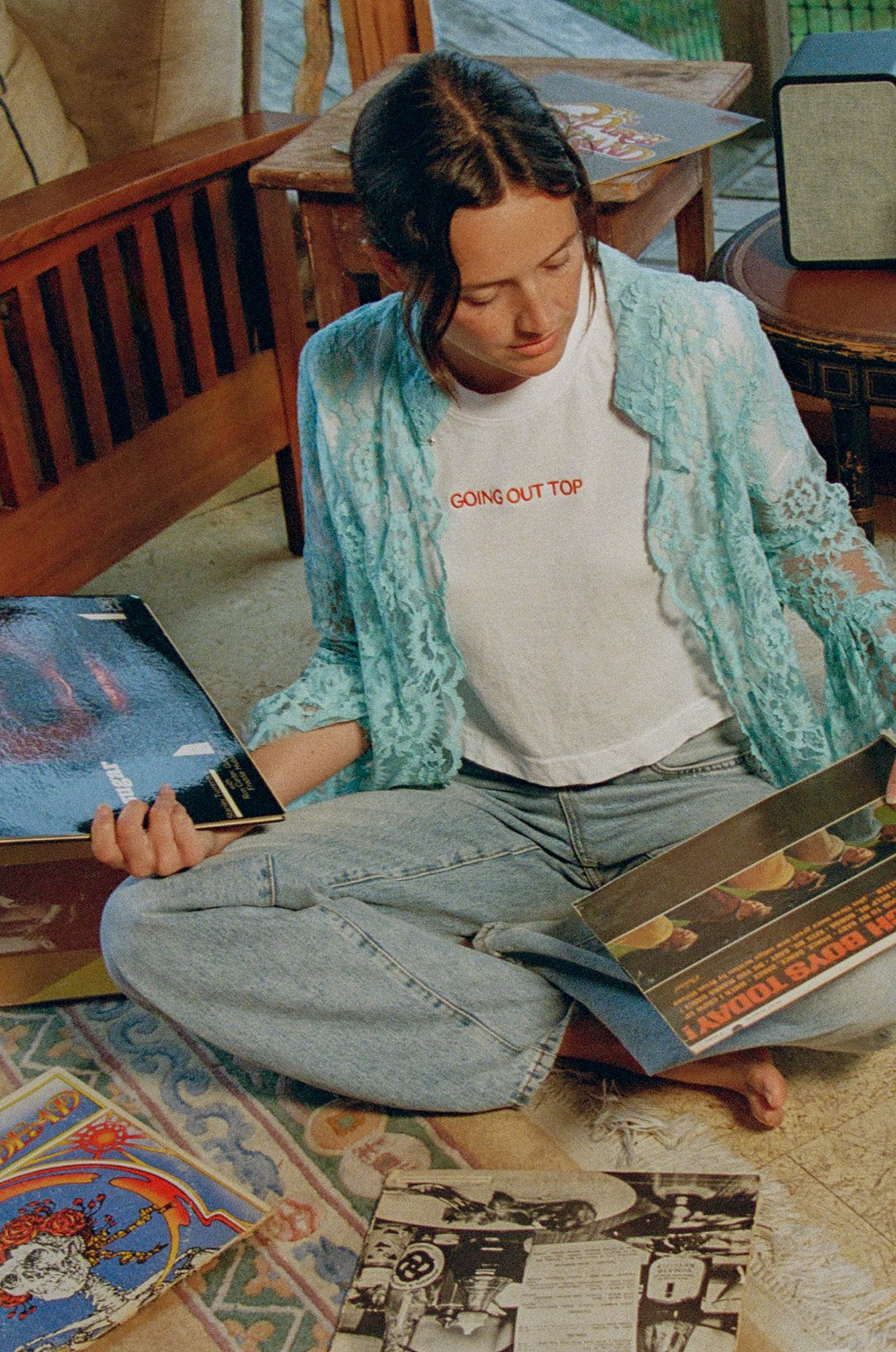 Model sitting cross-legged on the floor looking through records, wearing the Going Out Top with a lace shirt and jeans