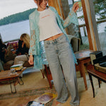 Model standing in a lakeside living room wearing the Going Out Top with a turquoise lace shirt and light-wash jeans