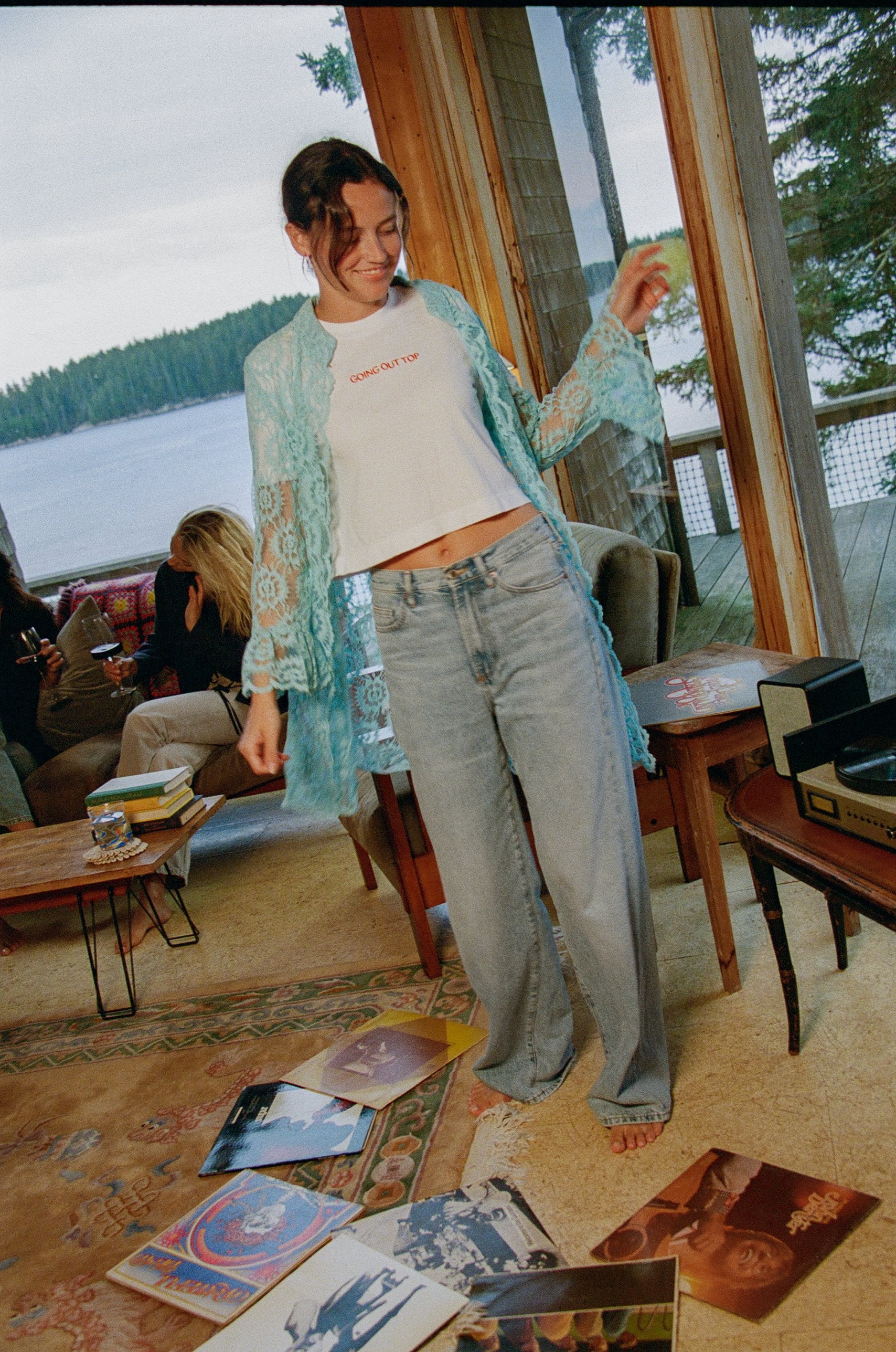 Model standing in a lakeside living room wearing the Going Out Top with a turquoise lace shirt and light-wash jeans