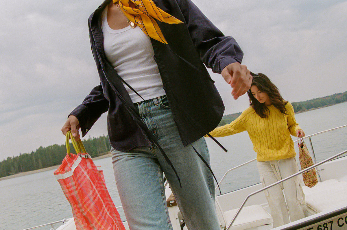 Two women on a boat, one wearing the By Dusk Two-Fer Top with light blue jeans and carrying a red patterned bag, while the other wears the Not Your Crewneck V-Neck Cable Knit Sweater in mustard yellow with white trousers.