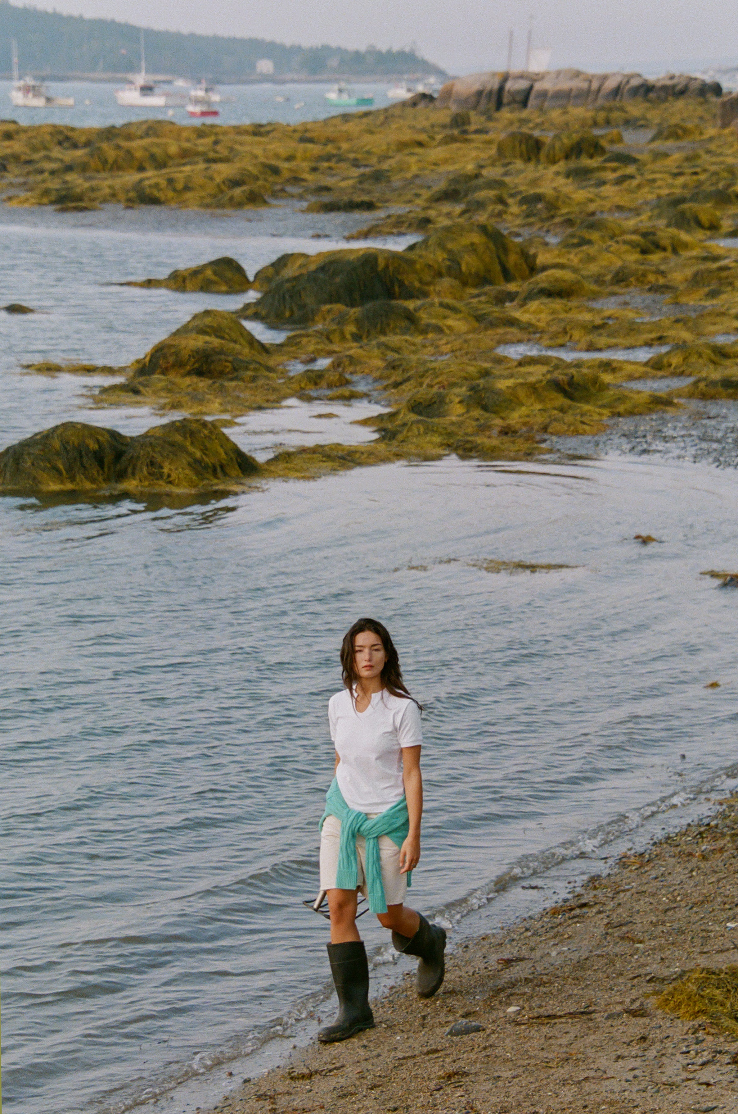 A woman walks along a rocky, seaweed-covered Maine shoreline wearing the By Dusk 70s Tee in white, rubber boots and a cable-knit sweater tied around her waist, with boats in the background.