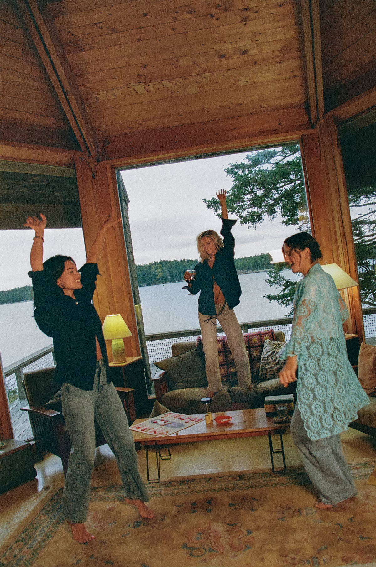 Three friends dancing in a wood-paneled lakeside cottage. One wears the By Dusk Casual Friday Button Down in black, another wears the Two-Fer Top, and the third wears a white T-shirt under a turquoise crochet duster.