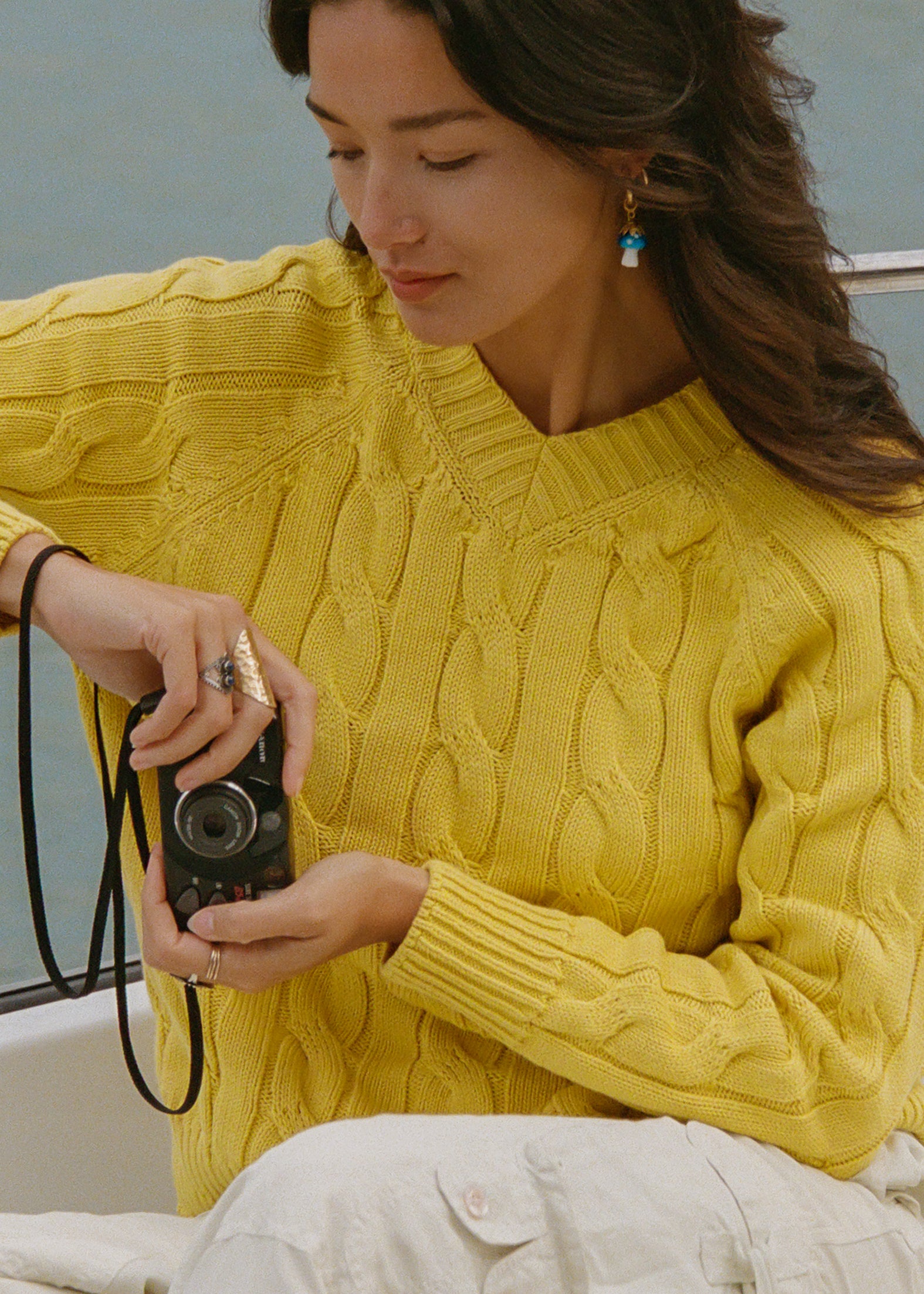 A woman wearing a mustard yellow not your crew neck cable-knit sweater and white pants, holding a vintage camera. She is wearing turquoise earrings and rings, with a soft-focus background of water and a boat.