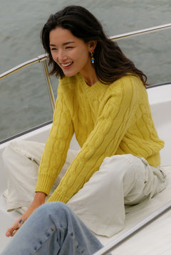 A woman sits on a boat wearing a mustard-yellow cable-knit crewneck sweater and white pants, with water in the background.