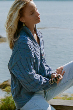 A woman sitting outdoors by the water wearing an off-duty quarter-zip in blue and light-wash jeans, holding a small camera.