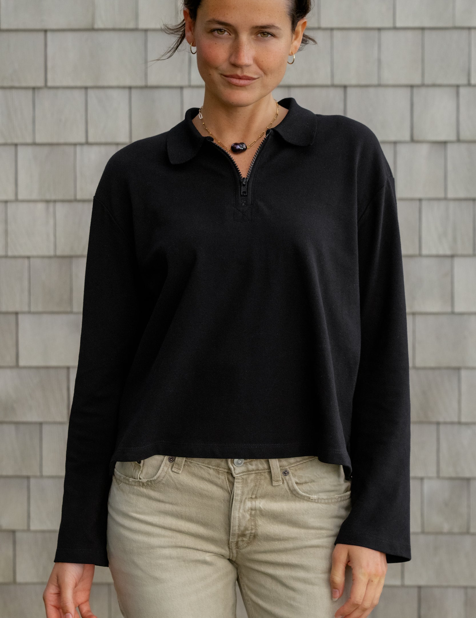 A woman wearing a long sleeve sunday funday polo in black and light beige jeans, standing against a gray shingled wall.