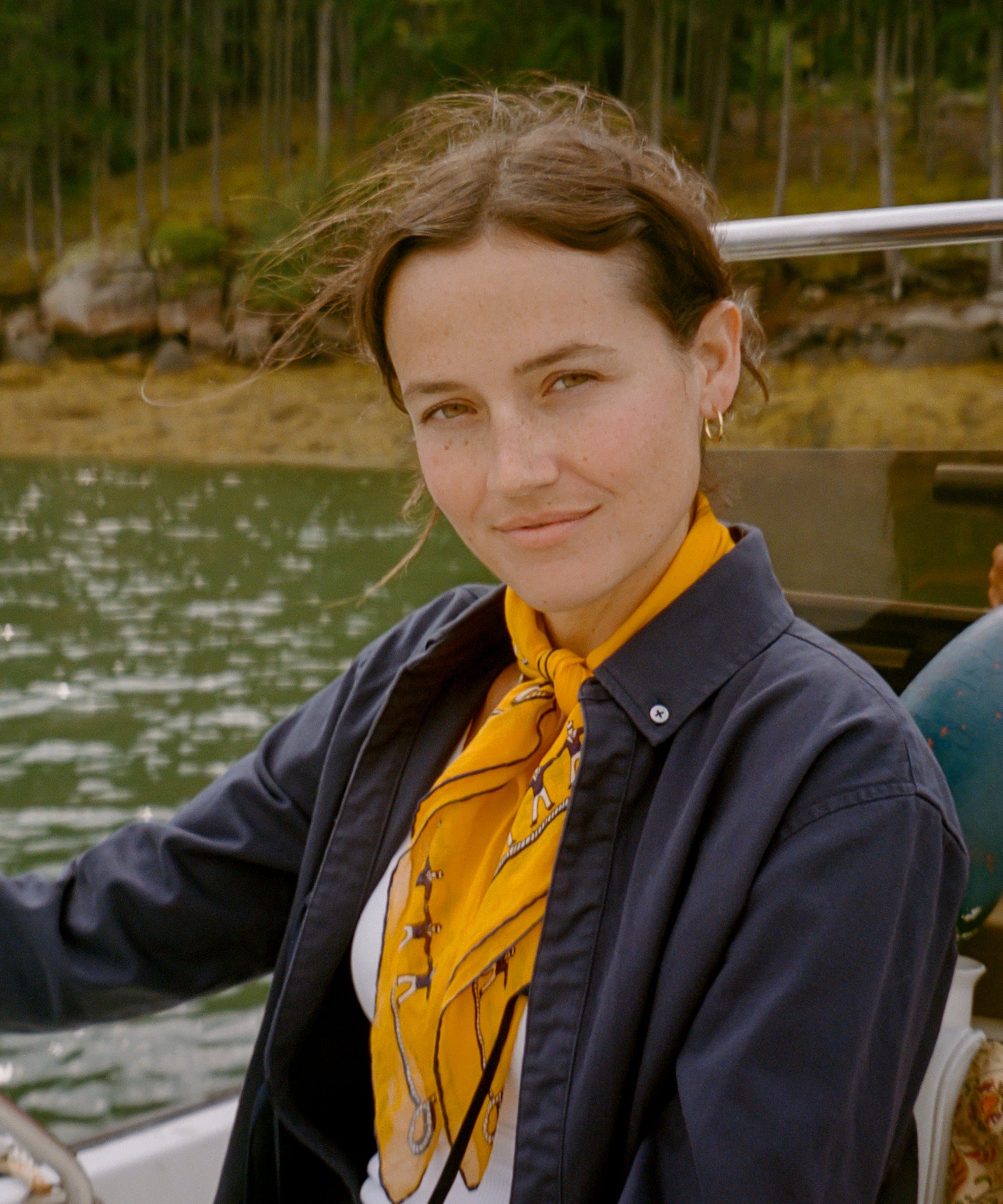 A woman wearing a navy twofer top shirt and a bright yellow patterned neck scarf, sitting on a boat with water and a forest in the background.