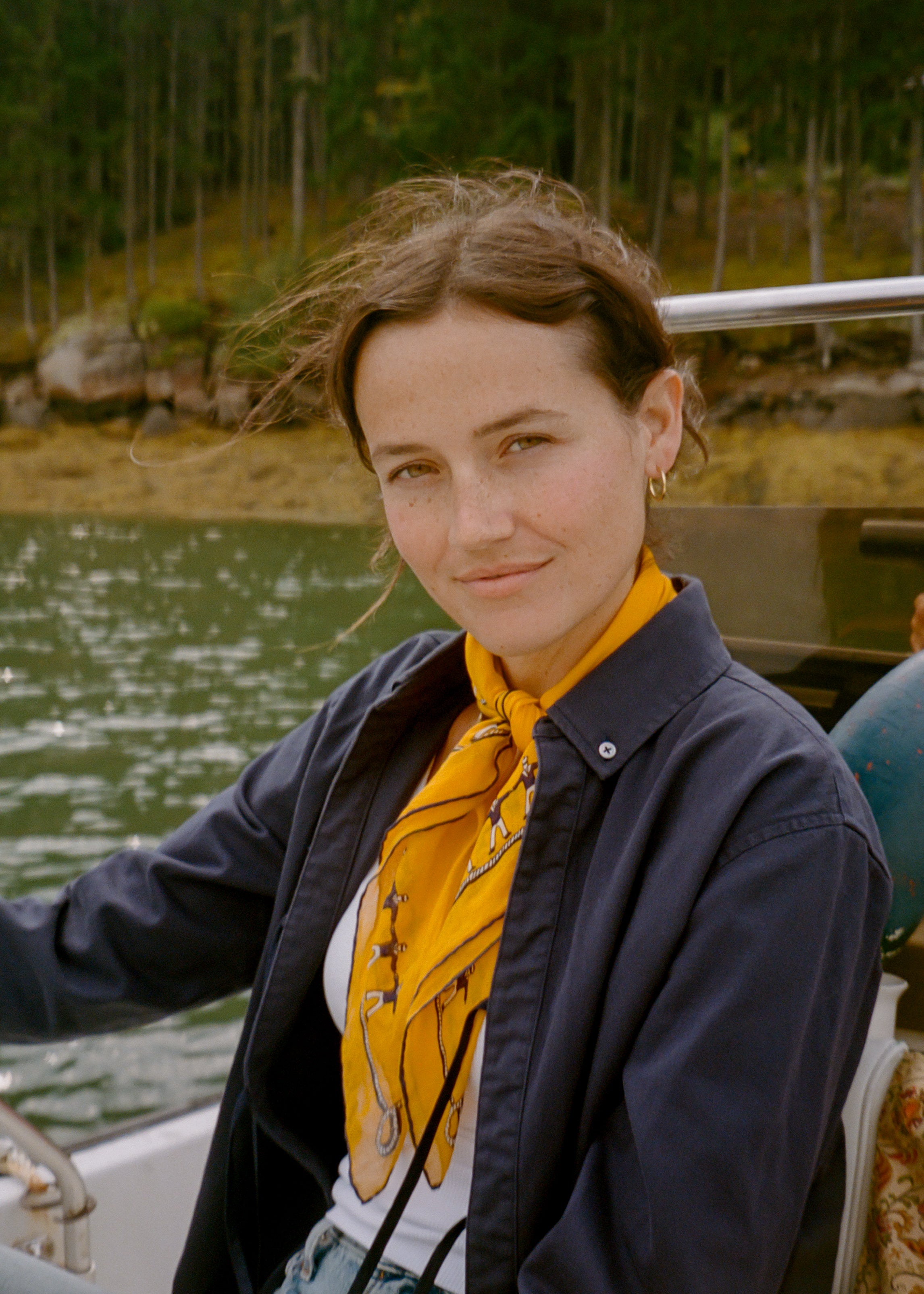 A woman wearing a navy twofer top shirt and a bright yellow patterned neck scarf, sitting on a boat with water and a forest in the background.