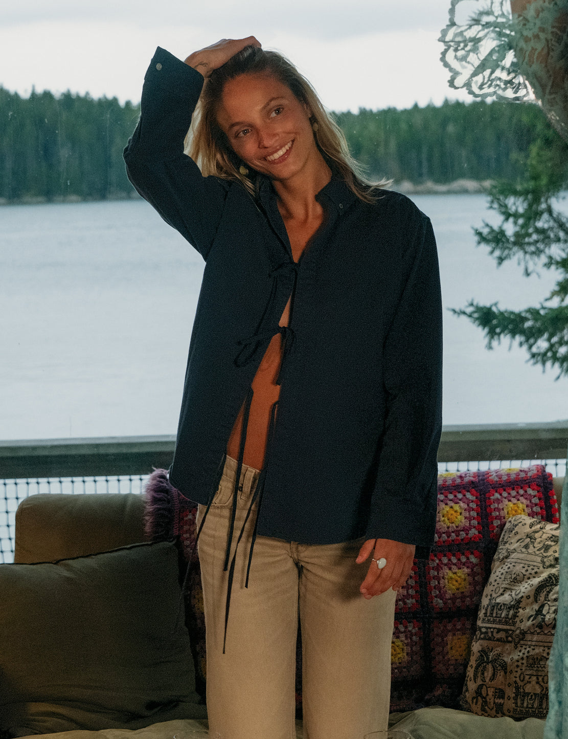 A woman stands indoors by a window with a ocean and forest view behind her, wearing a navy twofer top shirt with a front tie detail and beige pants.