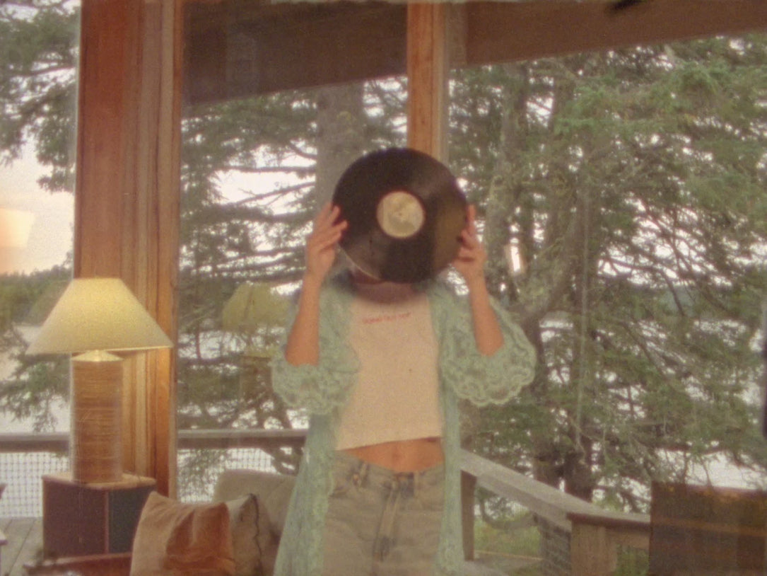 A woman holds a vinyl record over her face, wearing the Going Out Top Tee layered under a turquoise crochet duster with light-wash jeans.